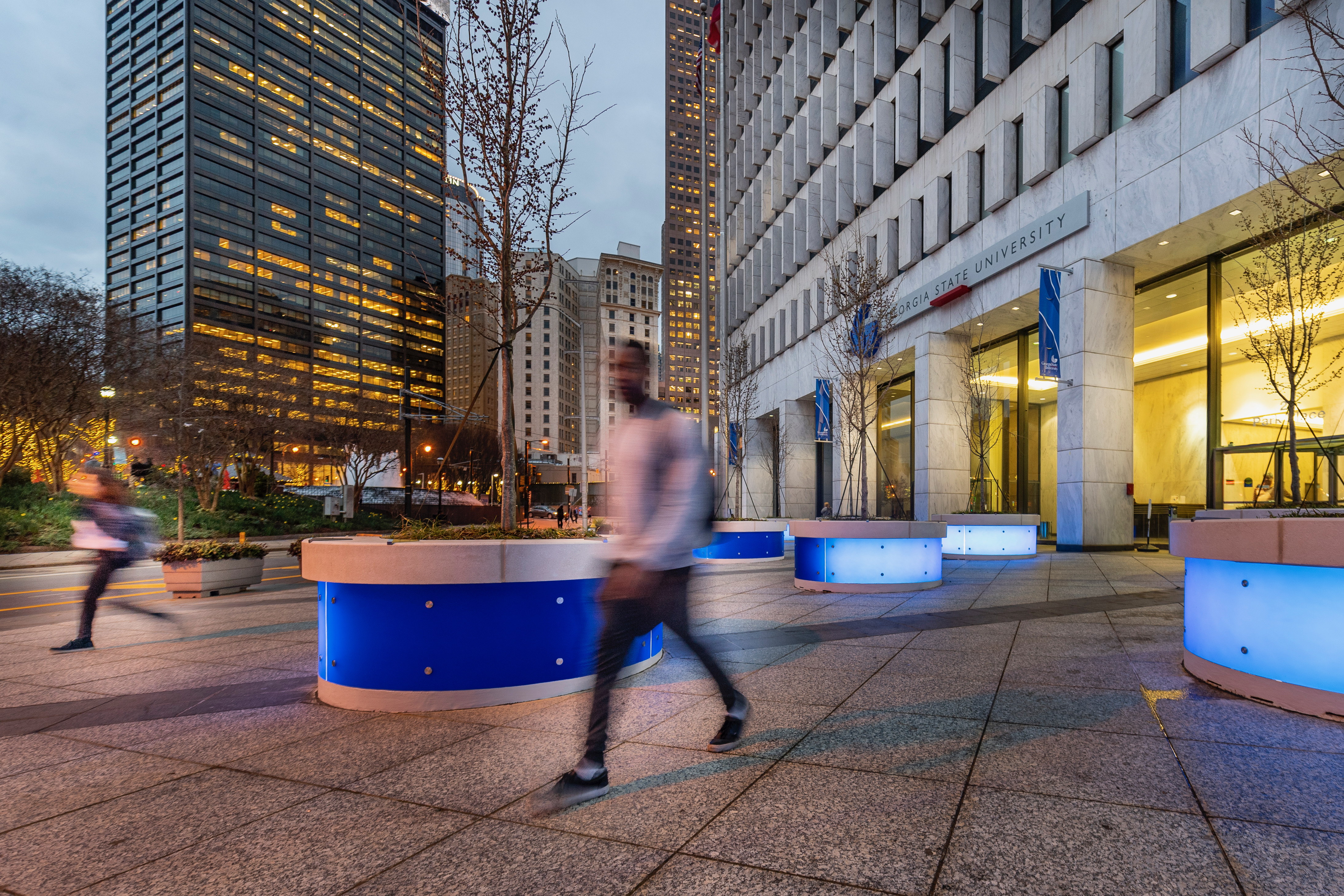 Students walking past a Georgia State University building entrance on a downtown Atlanta plaza at dusk, blue-lit planters lining the walkway